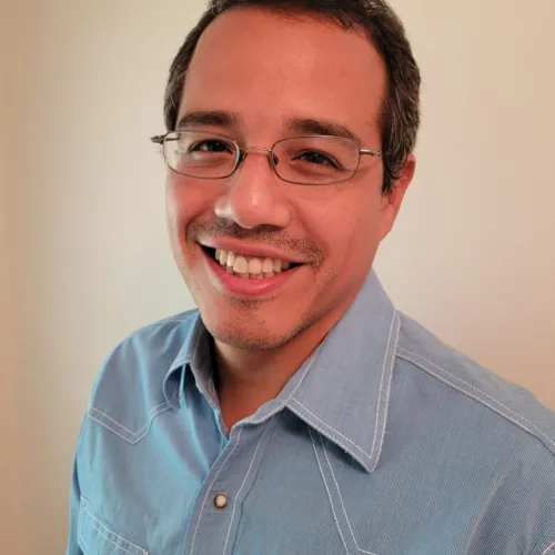 Professor Castillo, wears a blue shirt and glasses, and is smiling at the camera